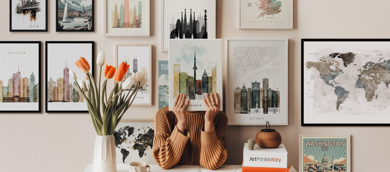 Person holding an art print in front of a wall displaying various framed cityscape artworks.