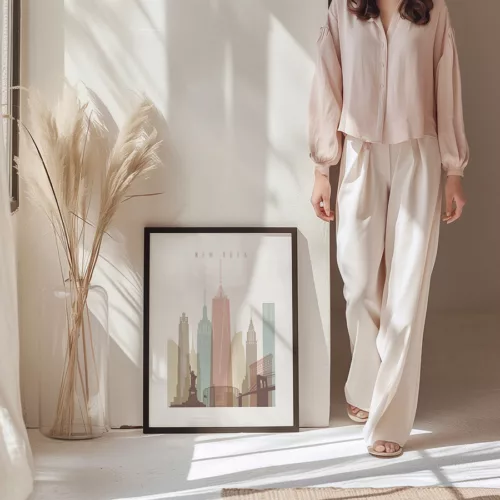 A model wearing a light blouse and wide-legged pants stands next to a framed artwork of New York City.
