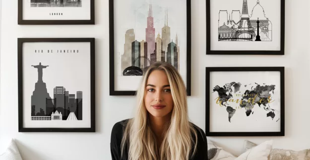 A woman with long blonde hair sitting in front of various framed cityscape artworks on a wall.