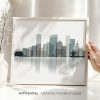A hand holding a framed minimalist cityscape art print featuring various buildings and a reflection.