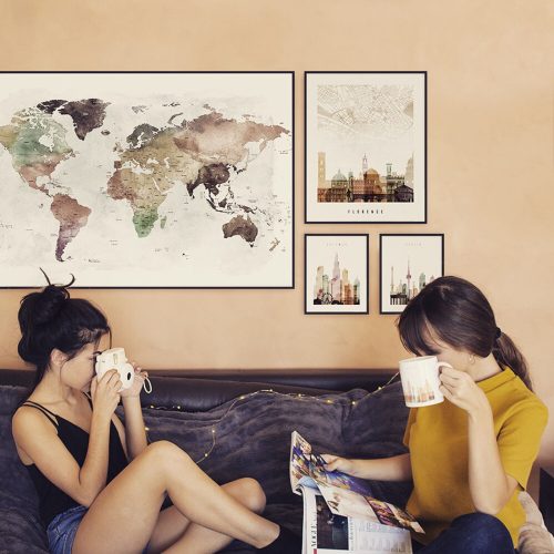 A world map and cityscape prints displayed on a wall with two women sitting on a couch, one holding a camera and the other a mug.