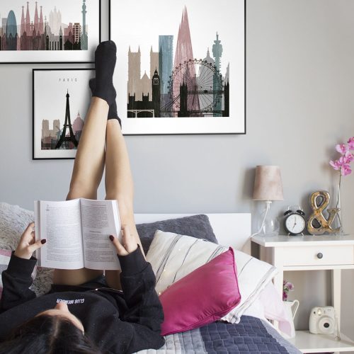 Individual reclining on a bed with legs up, reading a book in a room with framed cityscape art on the walls.