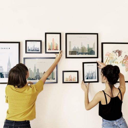 Two individuals arranging framed cityscape prints on a wall in an indoor setting.