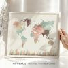 A watercolor world map featuring various colors representing different regions, held in a hand with a vase in the background.